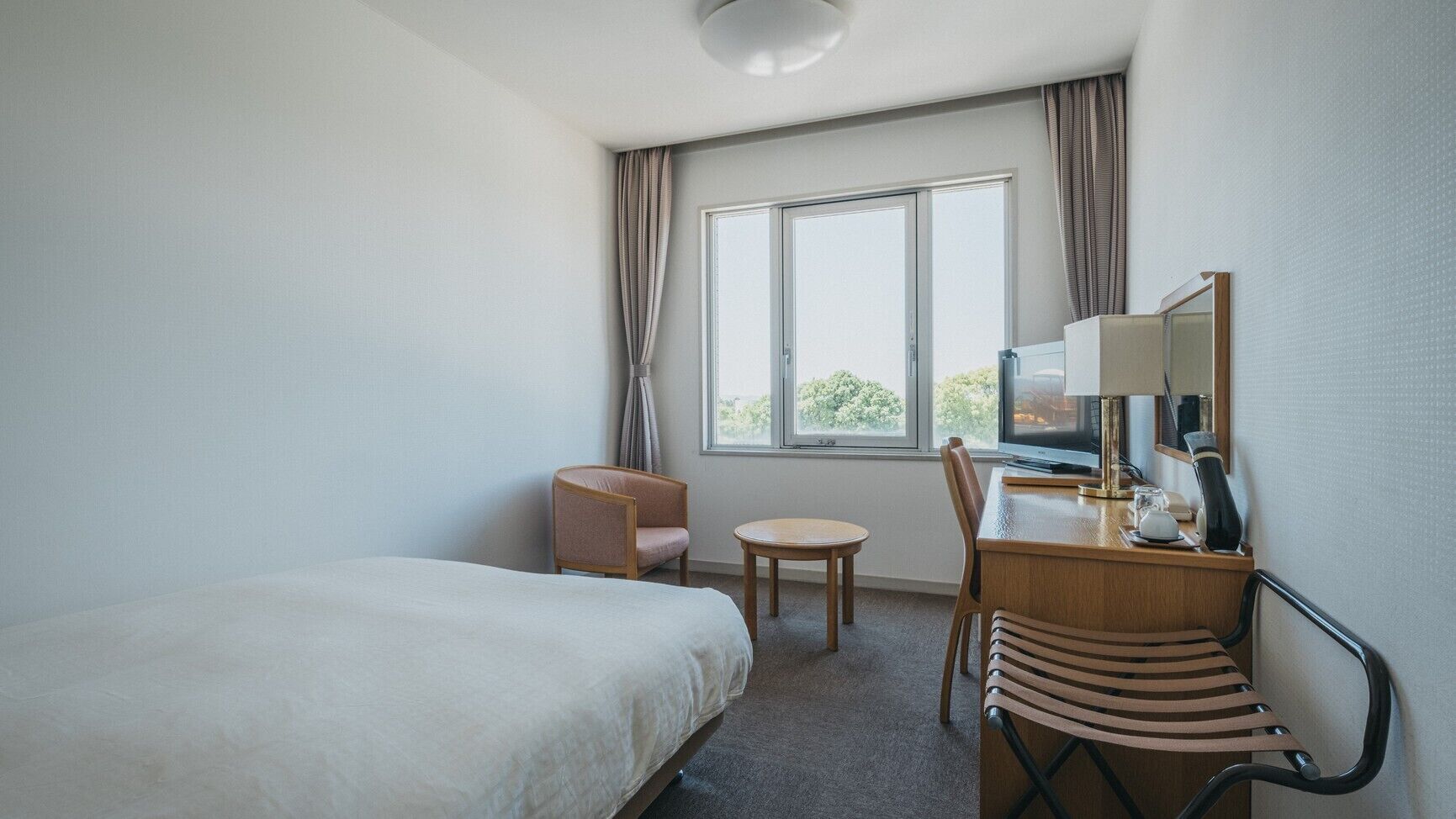 Ein Hotelzimmer mit einem Bett, einem Schreibtisch mit Stuhl, einem kleinen runden Tisch, einem Spiegel und einem Fenster mit Vorhängen.