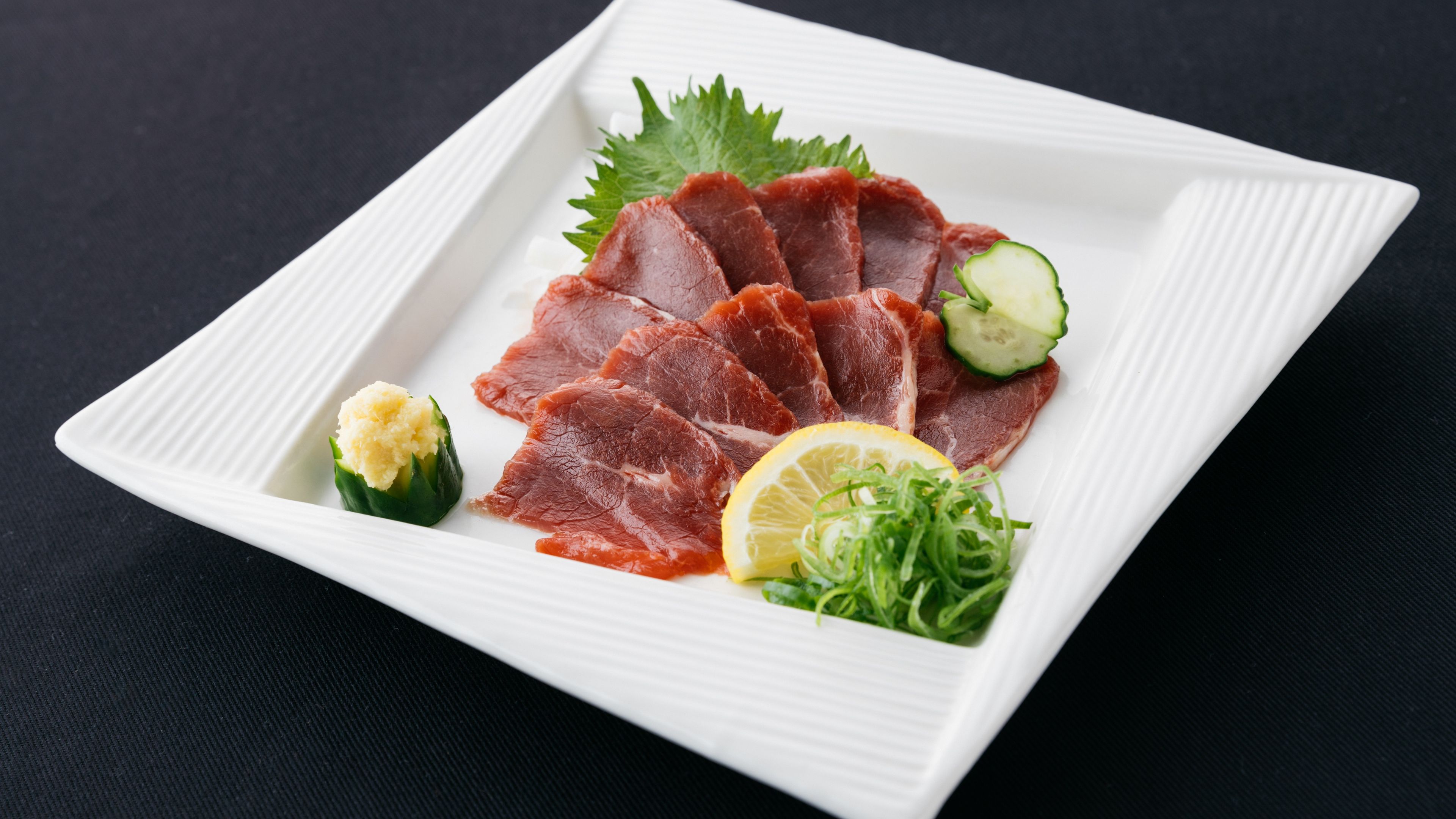 A white rectangular plate with thinly sliced red meat, a cucumber slice, a lemon wedge, and some green garnish.