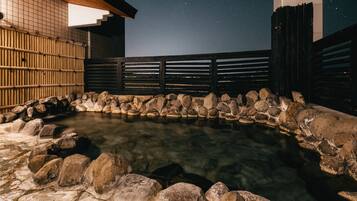 A stone hot spring surrounded by rocks and wooden fences, with a night sky visible above.