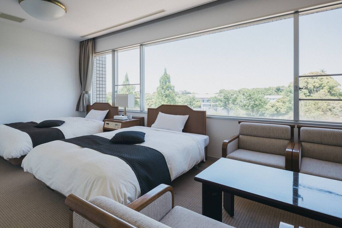 A hotel room with two beds, a sofa, a table, and large windows with a view of trees and buildings.
