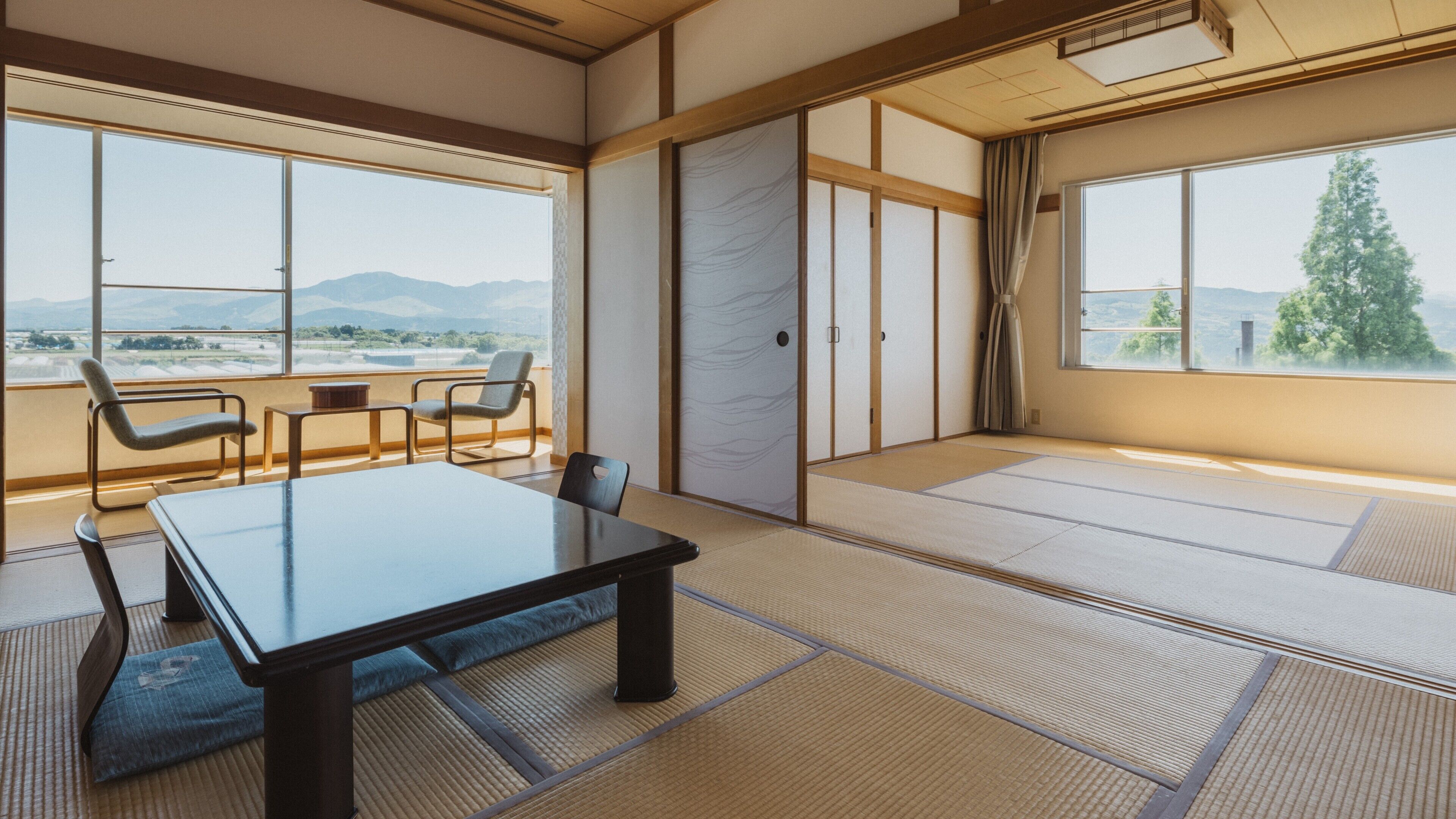 Ein Raum mit Tatami-Matten, einem niedrigen Tisch, Stühlen und großen Fenstern mit Blick auf Berge.