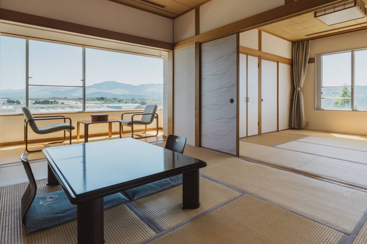 A tatami mat room with a low table, chairs, and large windows offering a view of mountains.