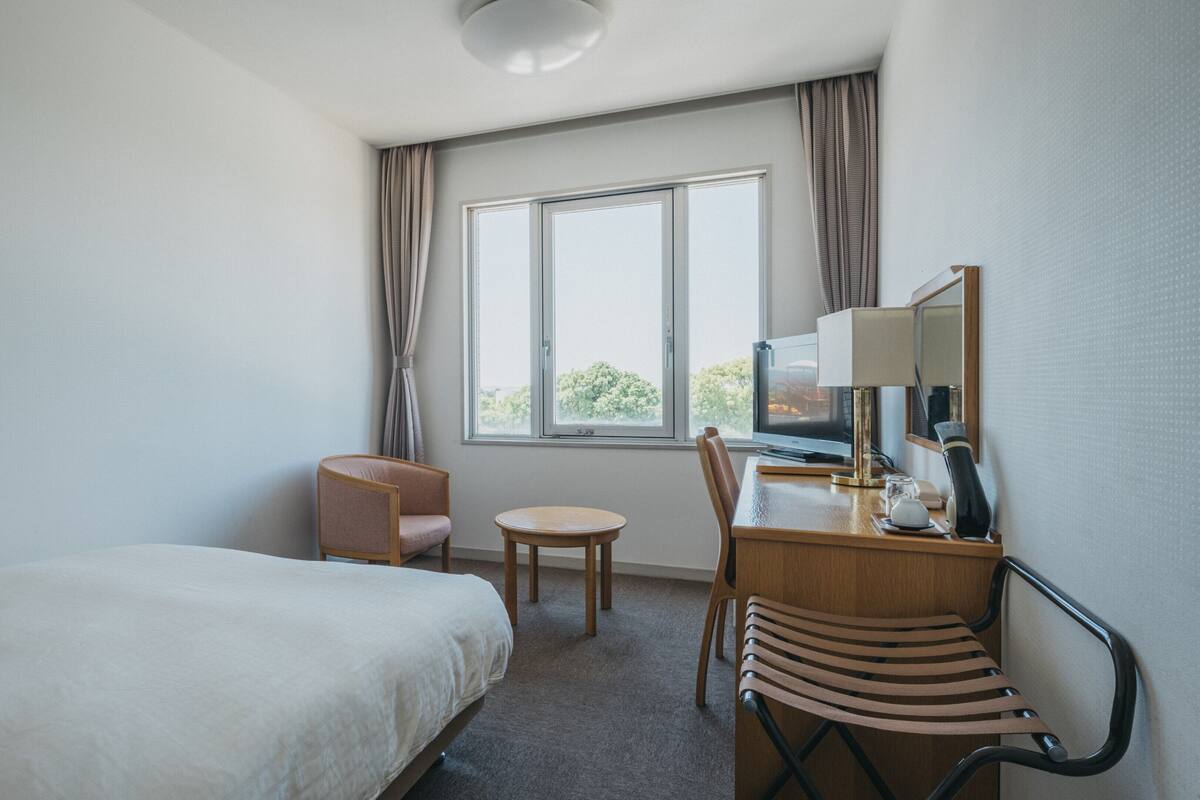 A hotel room with a bed, a desk with a chair, a small table, a lamp, a mirror, and a window with curtains.