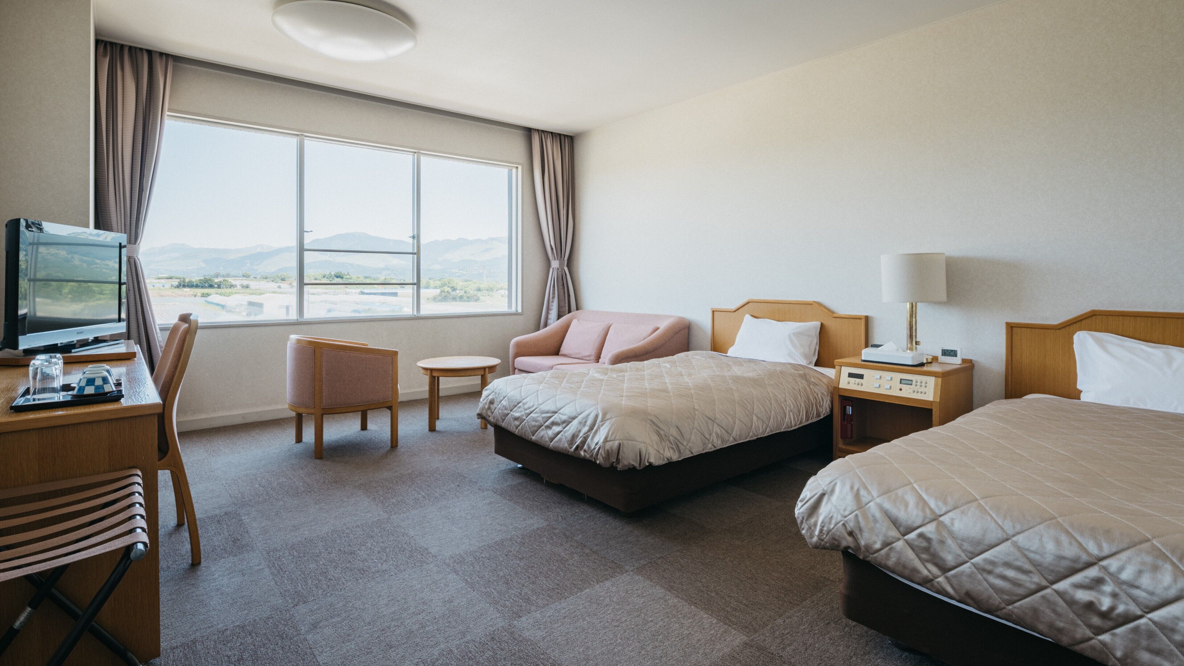 Ein Hotelzimmer mit Bett, Schreibtisch mit Computer, Stuhl, Nachttisch mit Lampe und Blick ins Freie.
