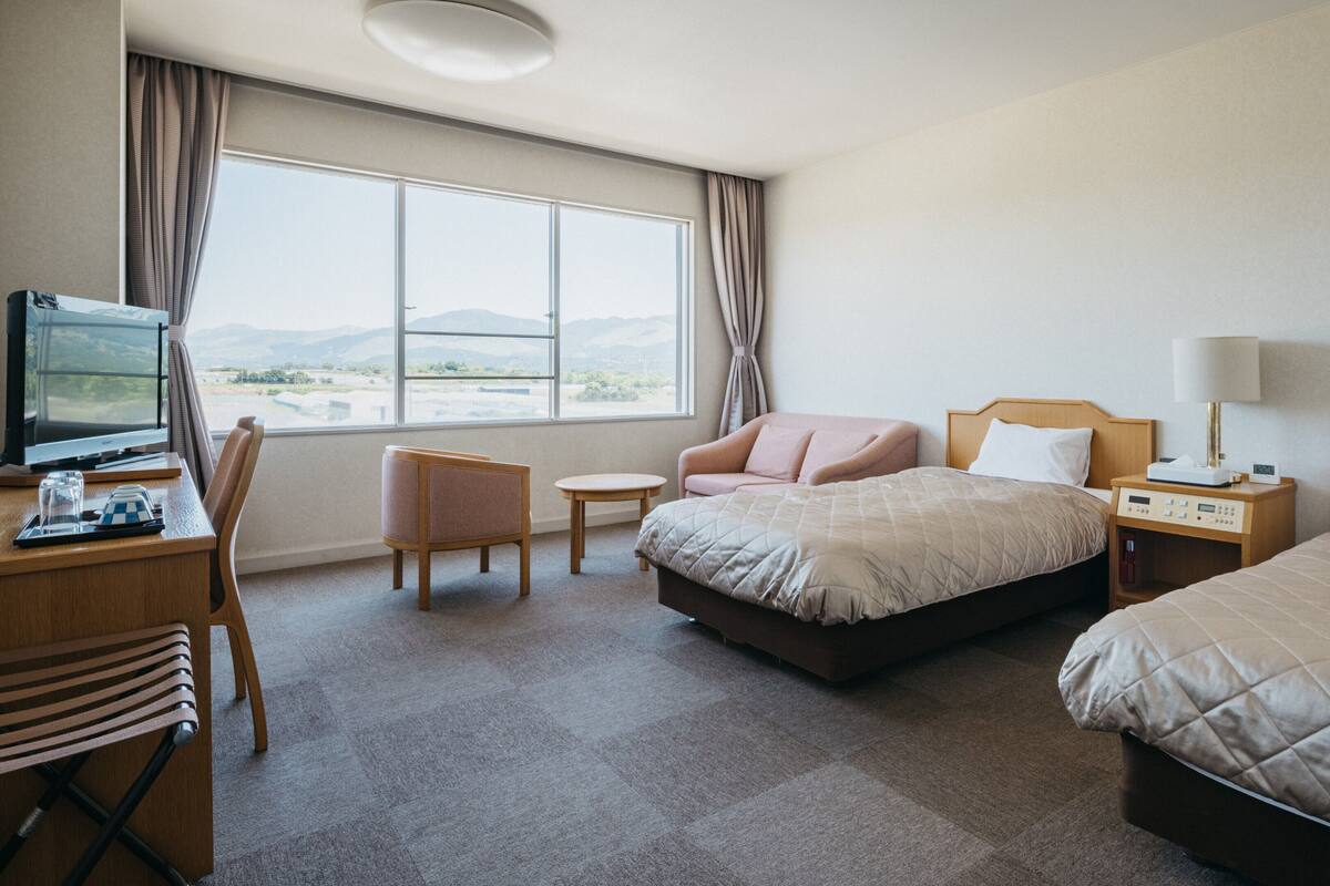 A hotel room with a bed, a desk with a computer, a chair, a bedside table with a lamp, and a view of the outdoors.