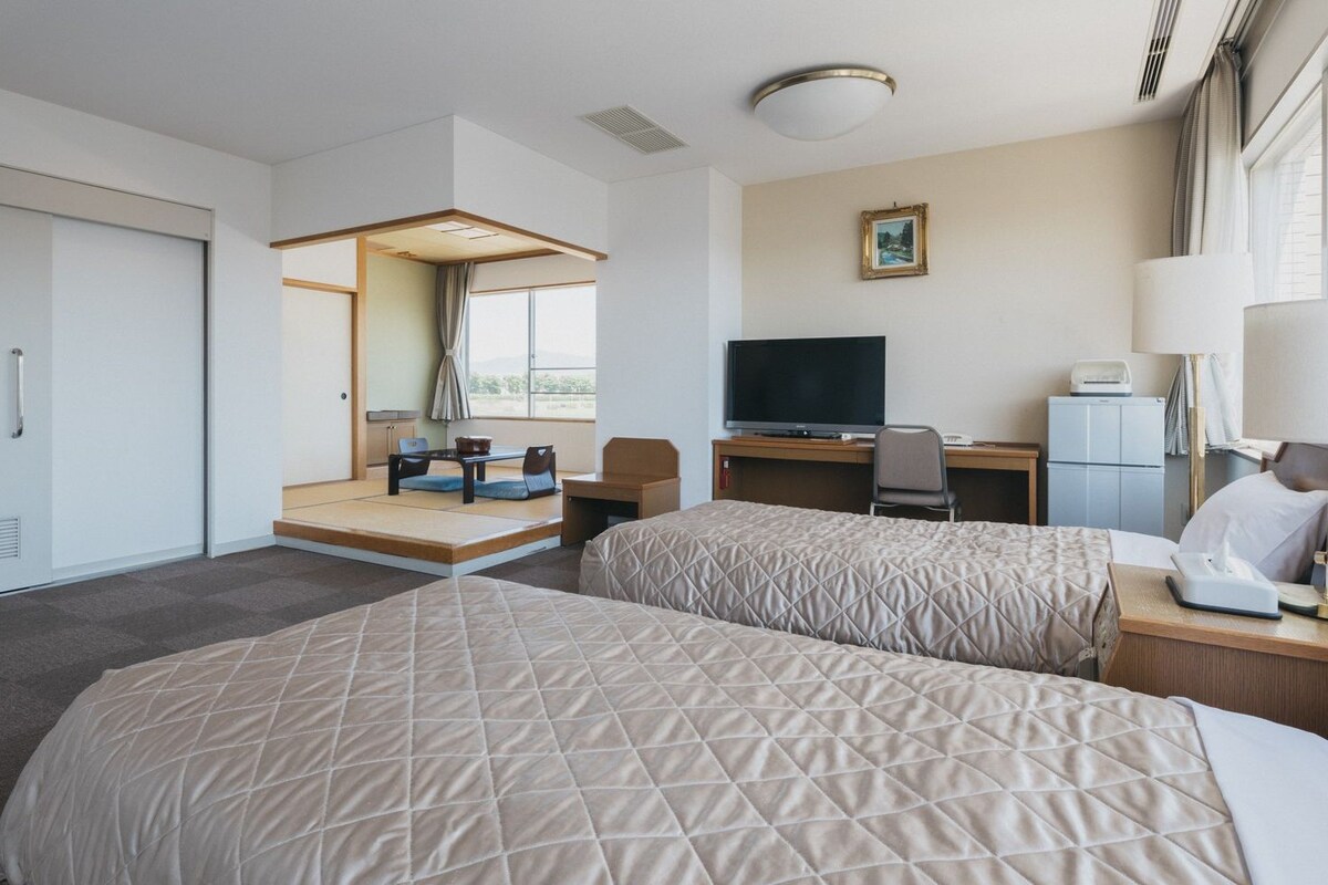 A hotel room with two beds, a desk with a TV, a chair, a lamp, and a window with a view.