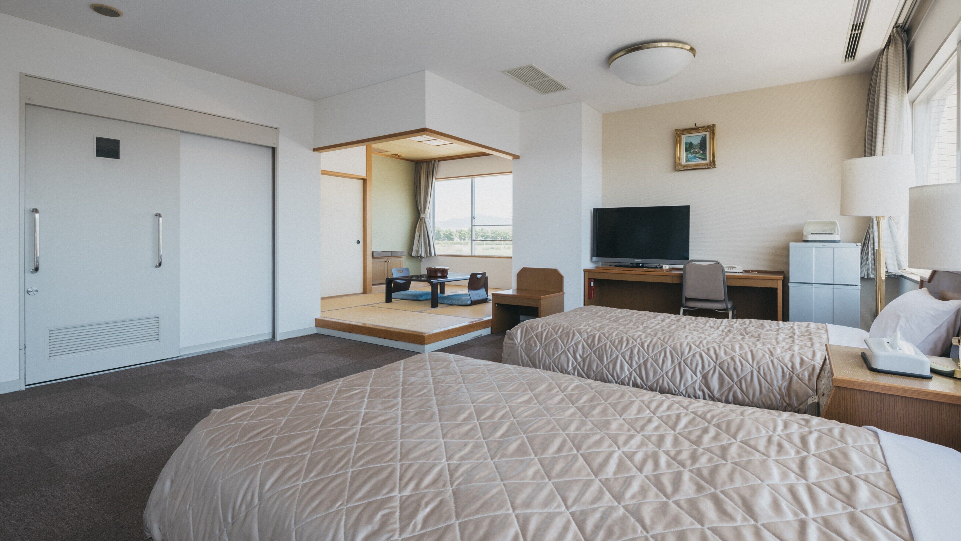 Ein Hotelzimmer mit zwei Betten, einem Schreibtisch mit Computer, einem Fernseher und einem kleinen Kühlschrank.