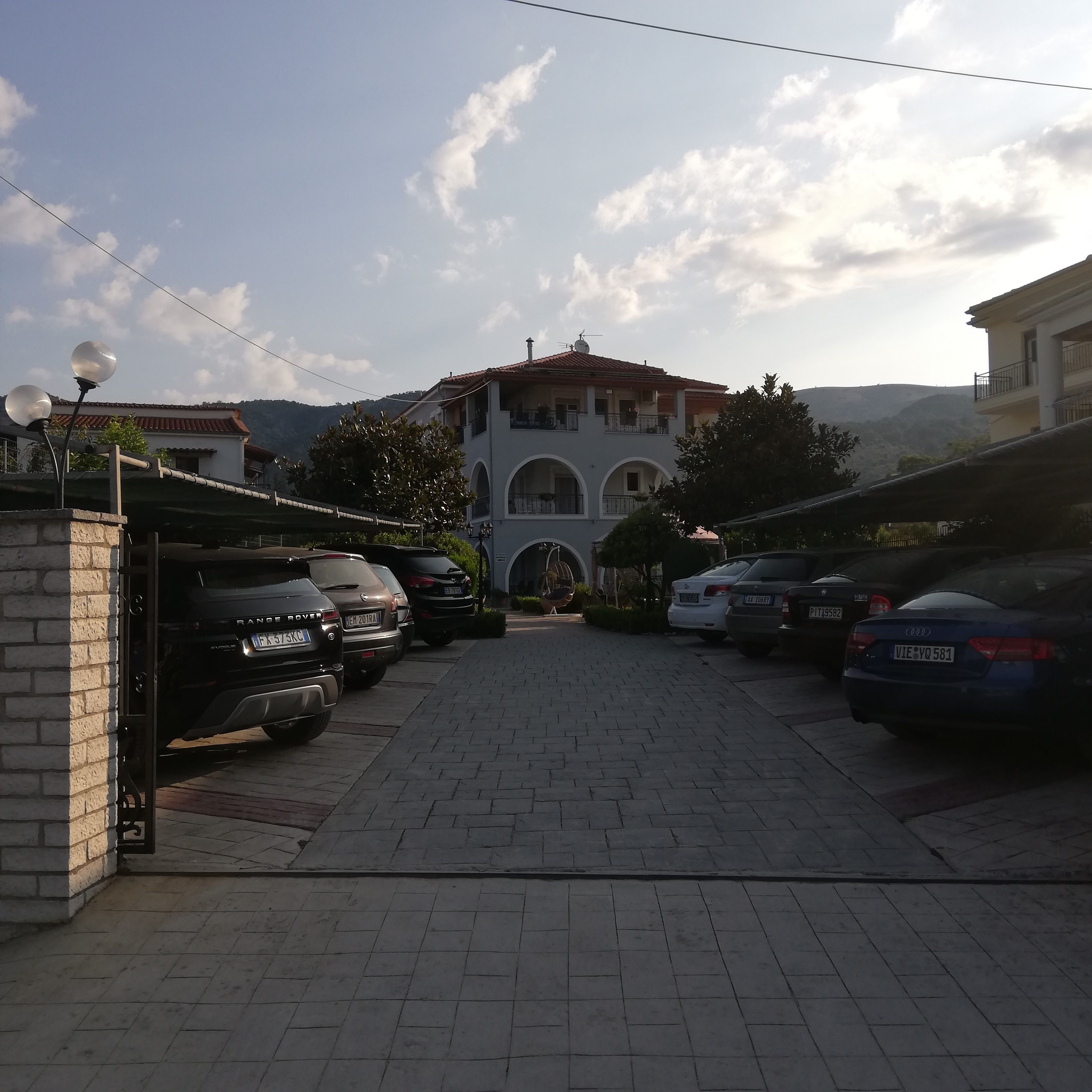 A parking area with multiple cars, a building with a red-tiled roof, and a mountainous backdrop.