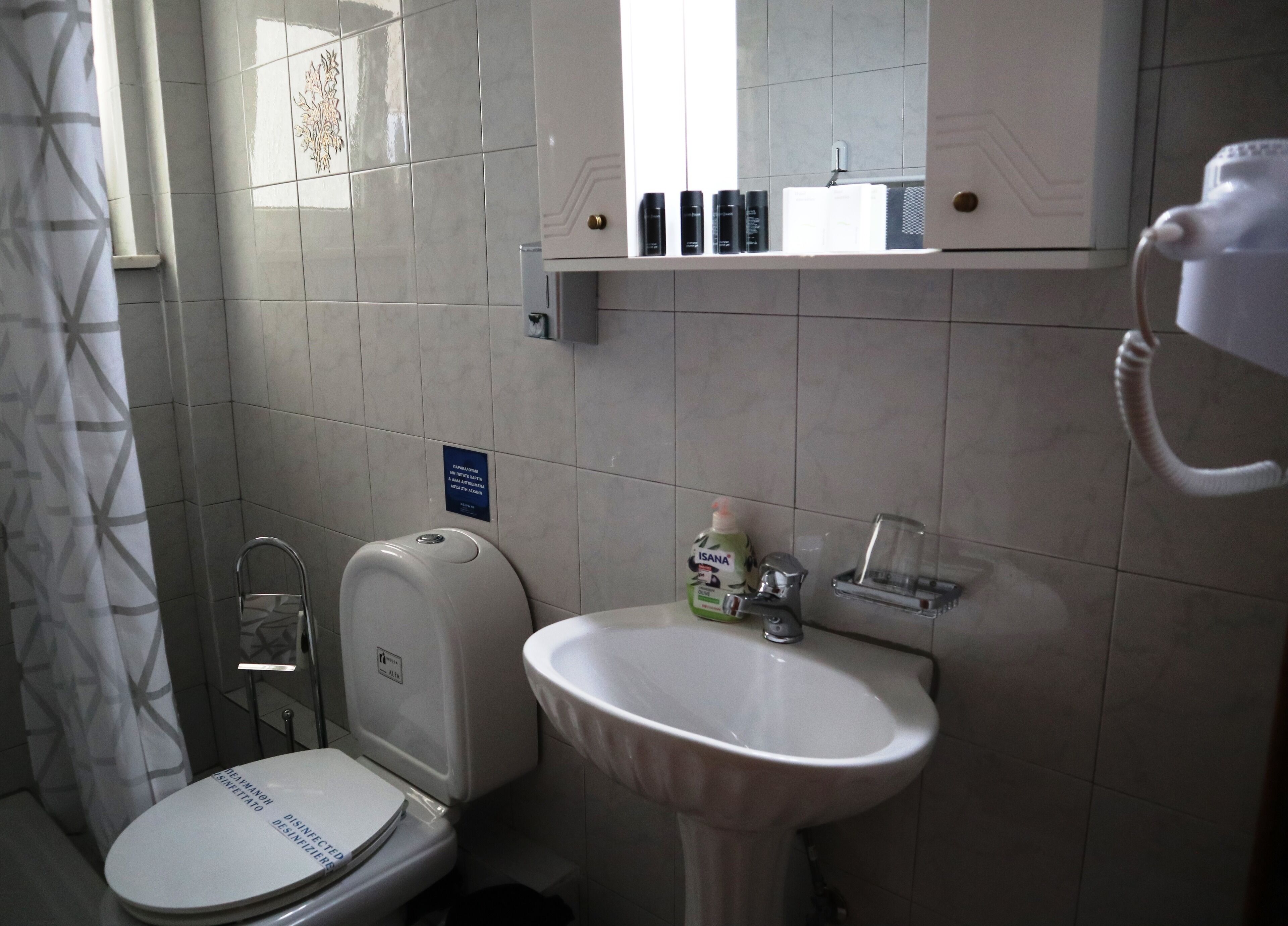 A bathroom with a toilet, sink, and hand dryer. There is a sign on the wall and a window with a curtain.