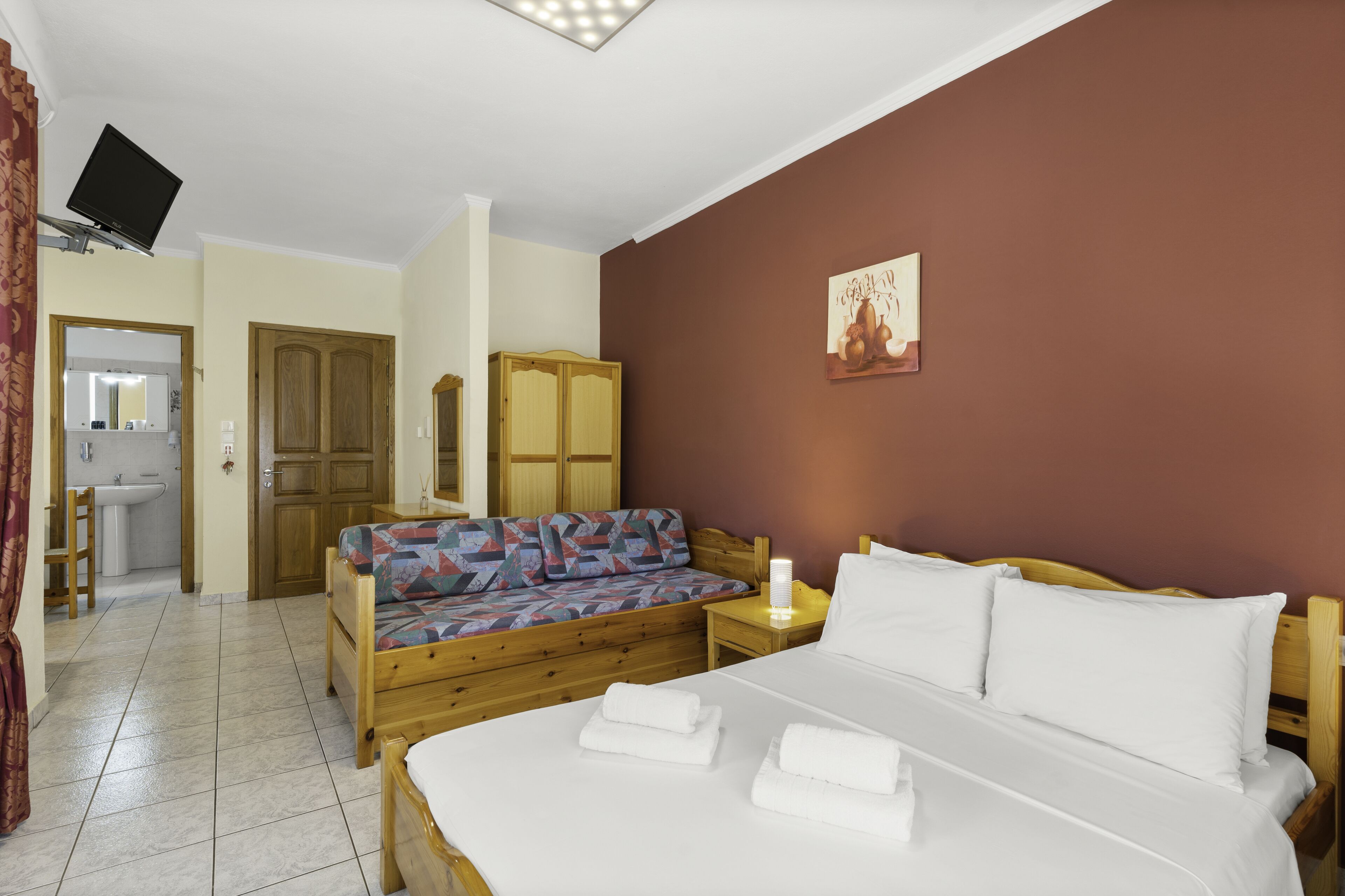 Une chambre à coucher comprenant un lit, un canapé, une armoire en bois, un téléviseur fixé au mur, et une salle de bain visible à travers une porte ouverte.