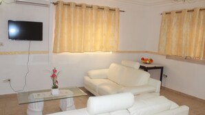 Un salón con un sofá blanco, una mesa de centro de cristal, un televisor de pantalla plana montado en la pared y una ventana con cortinas color beige.