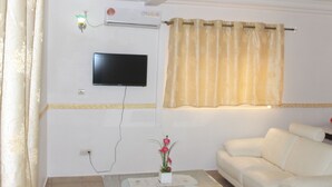 Una sala de estar con un sofá blanco, una mesa de centro de vidrio con un florero, un televisor de pantalla plana en la pared y una ventana con cortina.