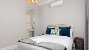 Una cama bien hecha con ropa blanca y almohadas decorativas, una mesita de noche con una lámpara y un aire acondicionado instalado en la pared.