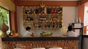 Zona de bar con pared de ladrillo, estantes llenos de botellas, una cesta de frutas y tres taburetes de bar.