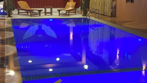 Una piscina con iluminación azul en su interior, sillas de lounge de madera y un fondo decorativo con figuras aladas.