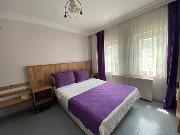 Un dormitorio con cabecera de madera, cama con manta morada, mesita de noche y ventana con cortinas traslúcidas.