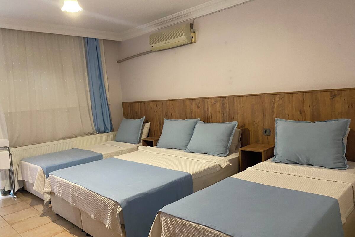 A hotel room with three single beds, each with a blue bedspread and white pillows. There is a wooden headboard, a window with blue curtains, and an air conditioning unit on the wall.