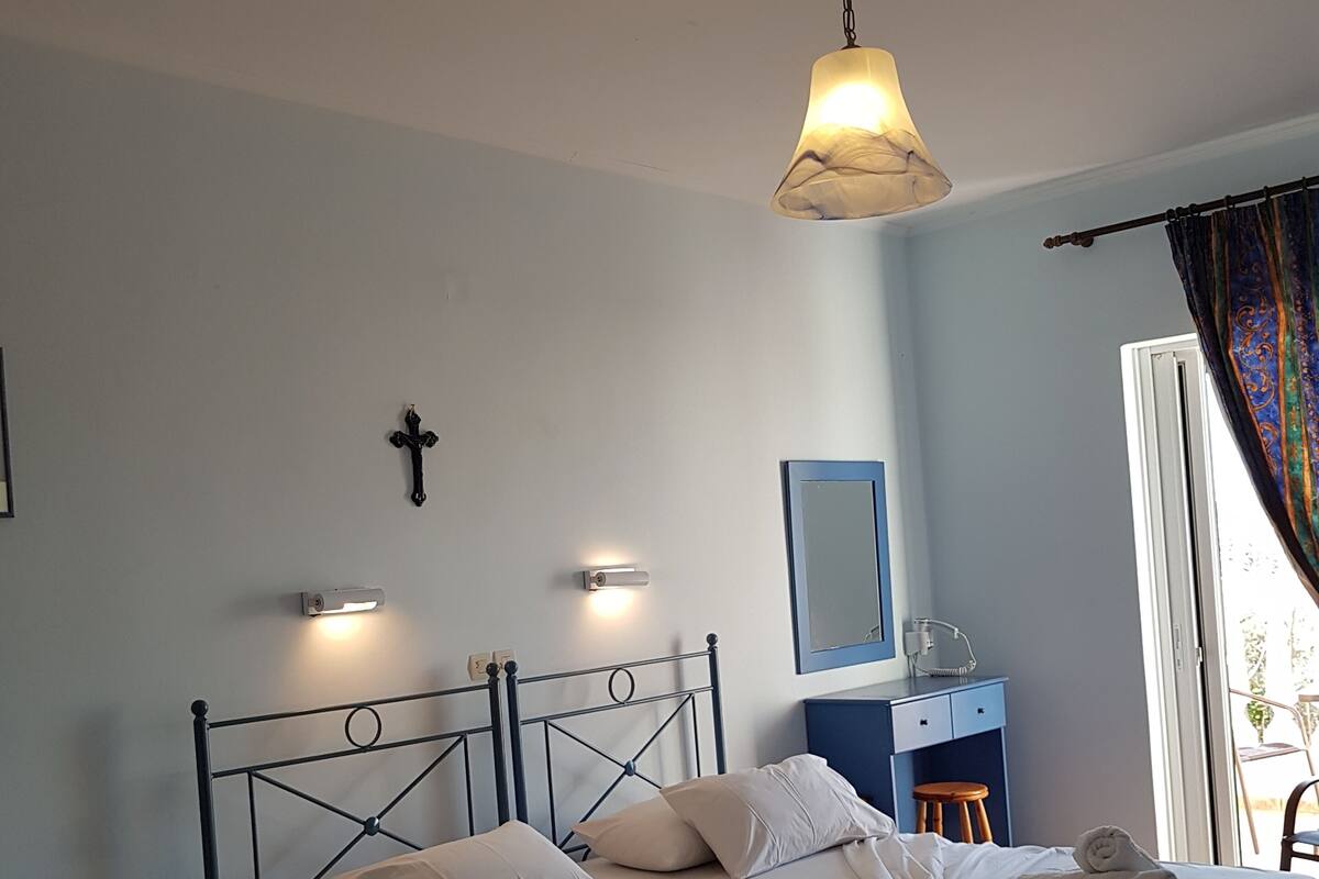 A neatly made bed with white linens and pillows, a bedside table, a desk with a chair, a cross on the wall, and a window with curtains.
