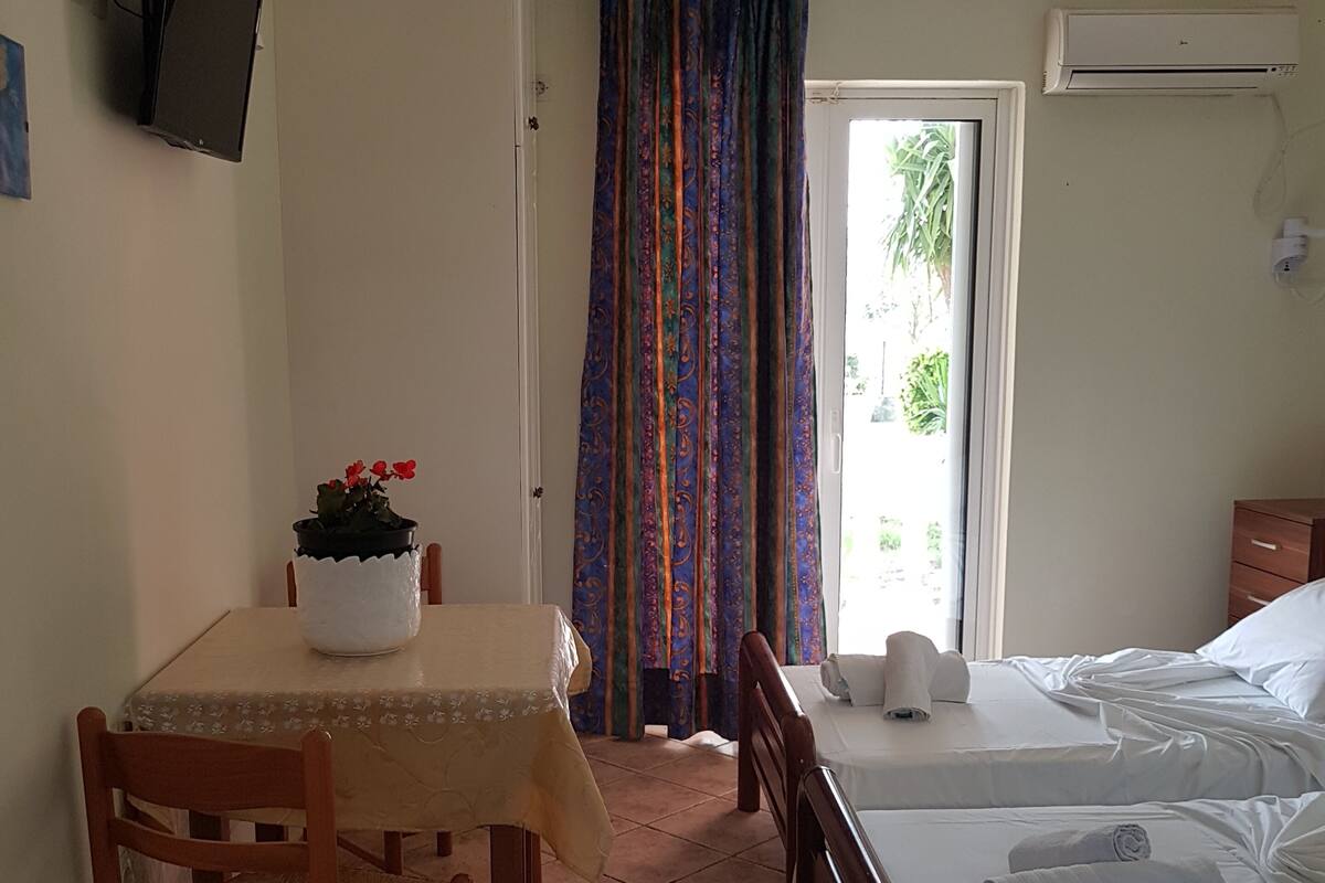 A small, tidy room with a bed, a table with a plant, a chair, and a TV mounted on the wall.