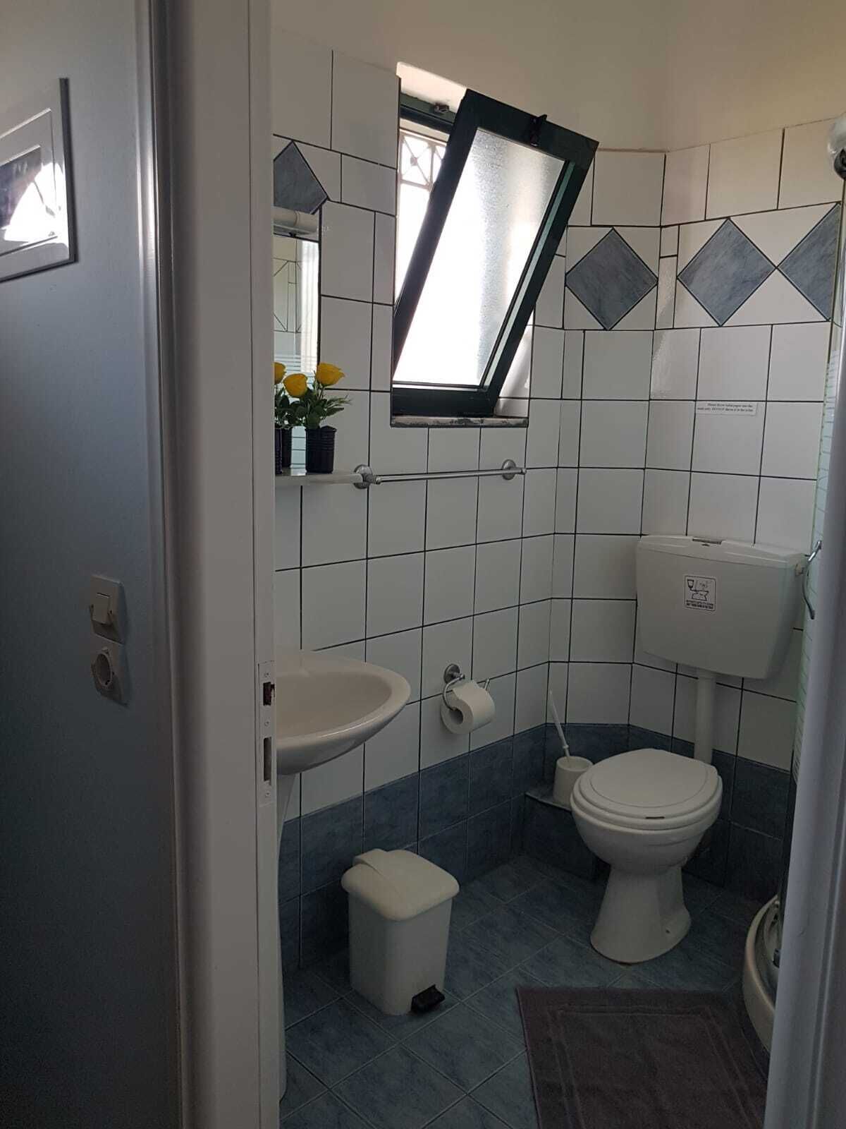 A compact bathroom with a window, sink, toilet, and a small trash bin.
