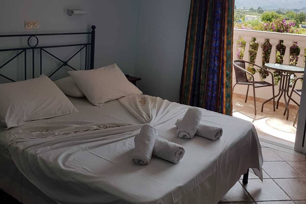 A neatly made bed with white linens and towels, a cross on the wall, and a balcony with a table and chairs.