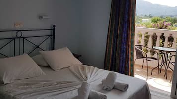 Cama bem arrumada com lençóis e toalhas brancos, uma cruz na parede e uma varanda com mesa e cadeiras.