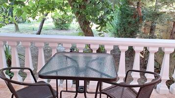Um pátio ao ar livre com mesa de vidro e cadeiras de vime, rodeado por um parapeito branco e vegetação exuberante.