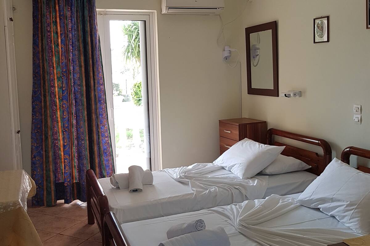 A hotel room with two beds, a wooden headboard, a small table, a chair, a window with curtains, an air conditioning unit, a mirror, and a wall-mounted phone.