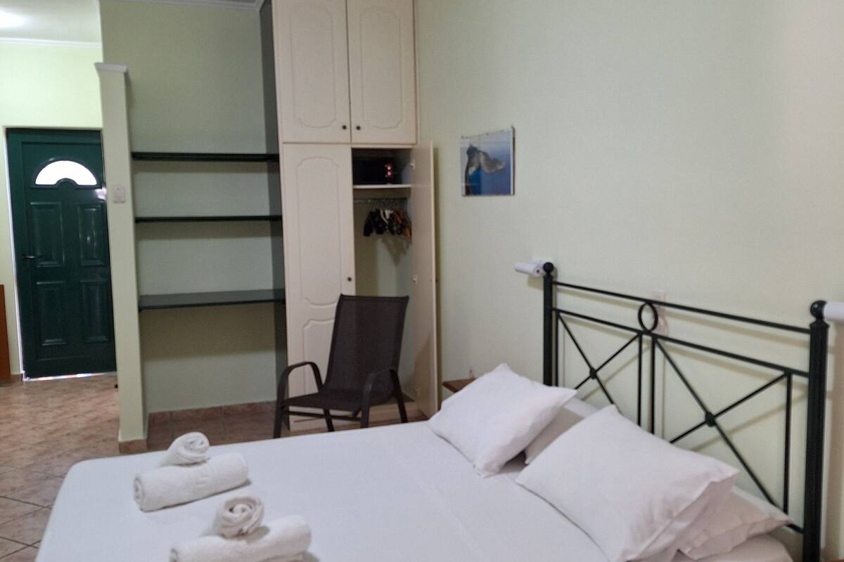 A neatly made bed with white linens and towels, a wooden nightstand, a metal headboard, a chair, and a wardrobe with open doors revealing hanging clothes.