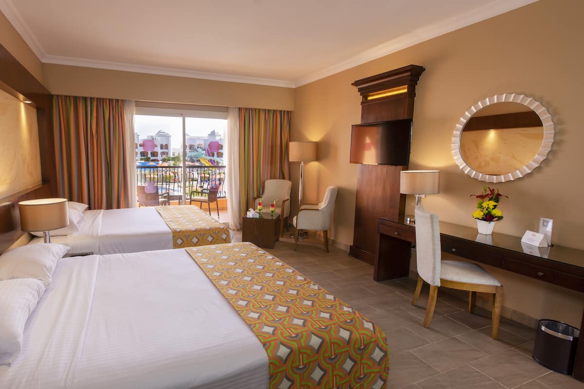 A hotel room with two beds, a desk, a chair, a mirror, and a view of the outside.