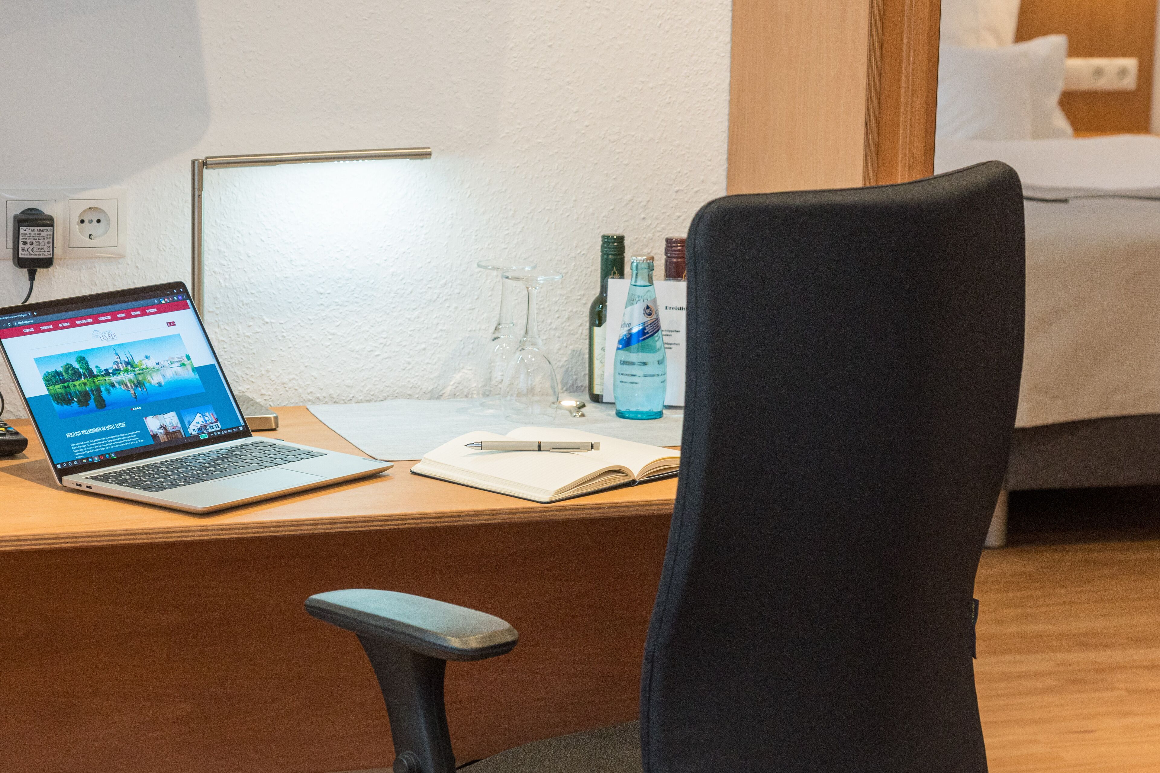 Une chambre d’hôtel avec un bureau en bois, un ordinateur portable, un carnet, un stylo, un verre et une bouteille d’eau.