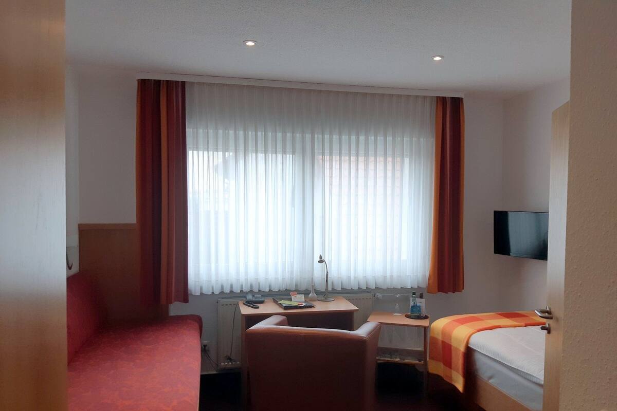 A hotel room with a bed, a desk, a chair, a TV, and a window with curtains.