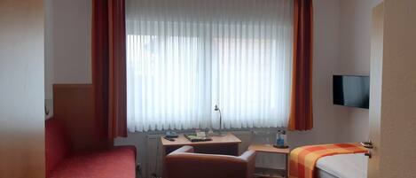 Habitación de hotel con cama, escritorio, silla, televisor y ventana con cortinas.