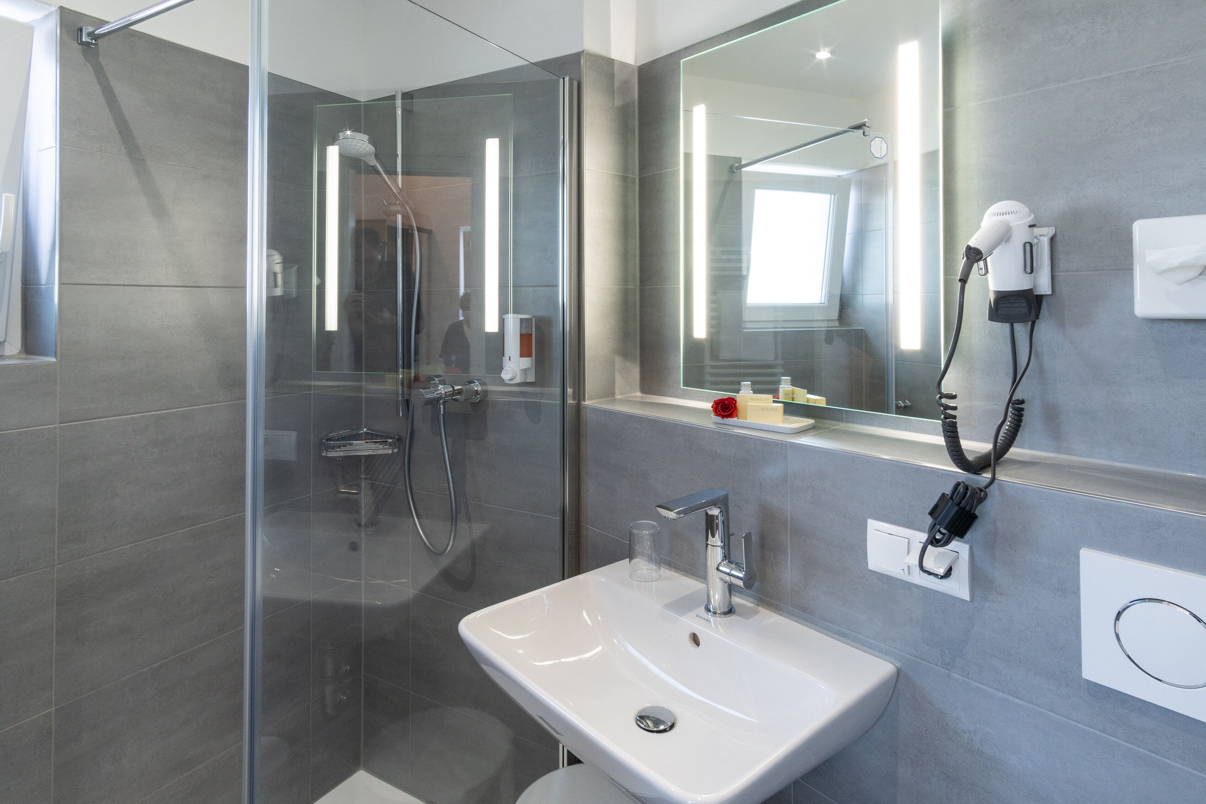 A modern bathroom with a glass-enclosed shower, a white sink, and a mirror with a hairdryer plugged in.