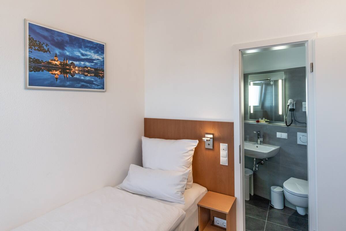 A hotel room with a bed, a nightstand, a wall-mounted light, a painting of a cityscape, and a bathroom with a sink and toilet.