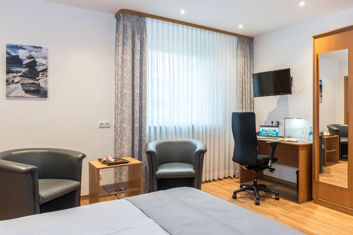 A hotel room with a bed, two chairs, a desk with a computer, a TV, and a window with curtains.