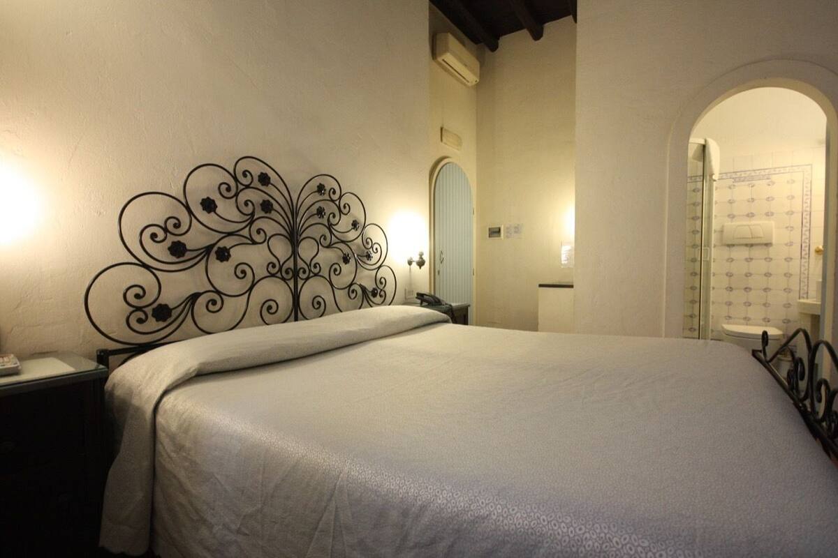 A bedroom with a bed, a nightstand, a decorative metal headboard, a bathroom with a shower and toilet visible through an archway.