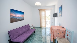 A room with a purple sofa, a small round table, a dining table with a patterned tablecloth, a white chair, and a TV on the wall. - Fotini Studios (Kos)