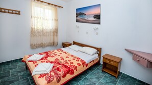 A bedroom with a bed, a nightstand, a wooden bench, a wall-mounted shelf, and a painting of a beach scene. - Fotini Studios (Kos)