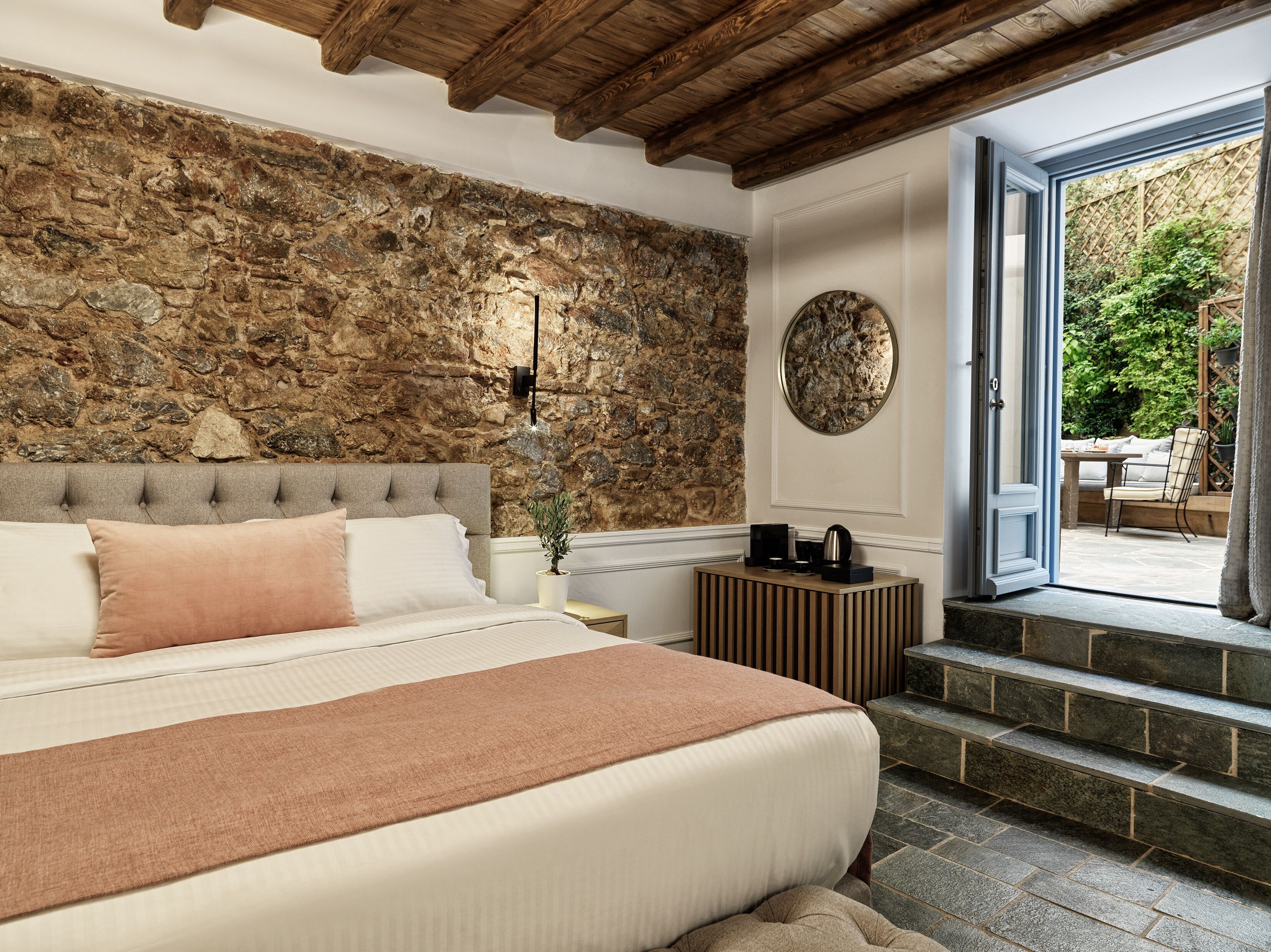 A bedroom with a stone wall, a bed with a headboard, a bedside table with a lamp, and a view of a garden through an open door.