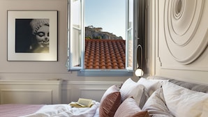 Un dormitori amb un llit, almoinos, una finestra que dona a un edifici, i un quadre al mur.