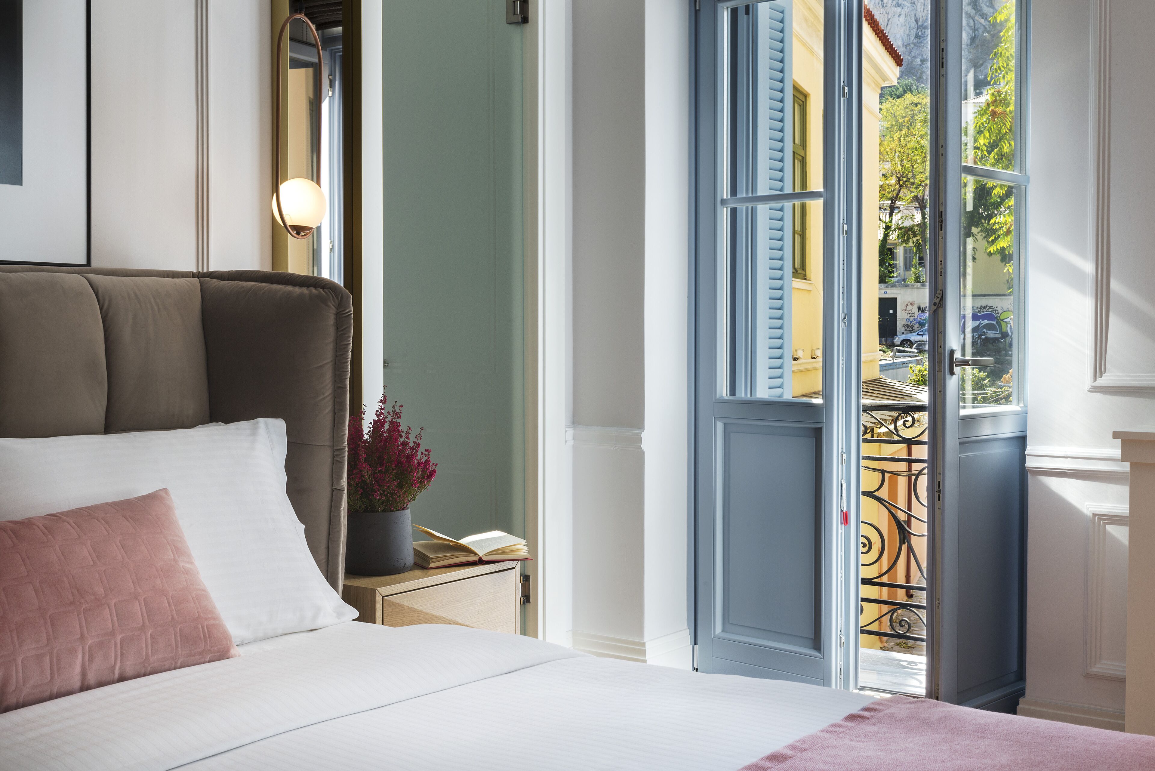 A bedroom with a bed, a nightstand, a potted plant, and a view through an open door to the outside.