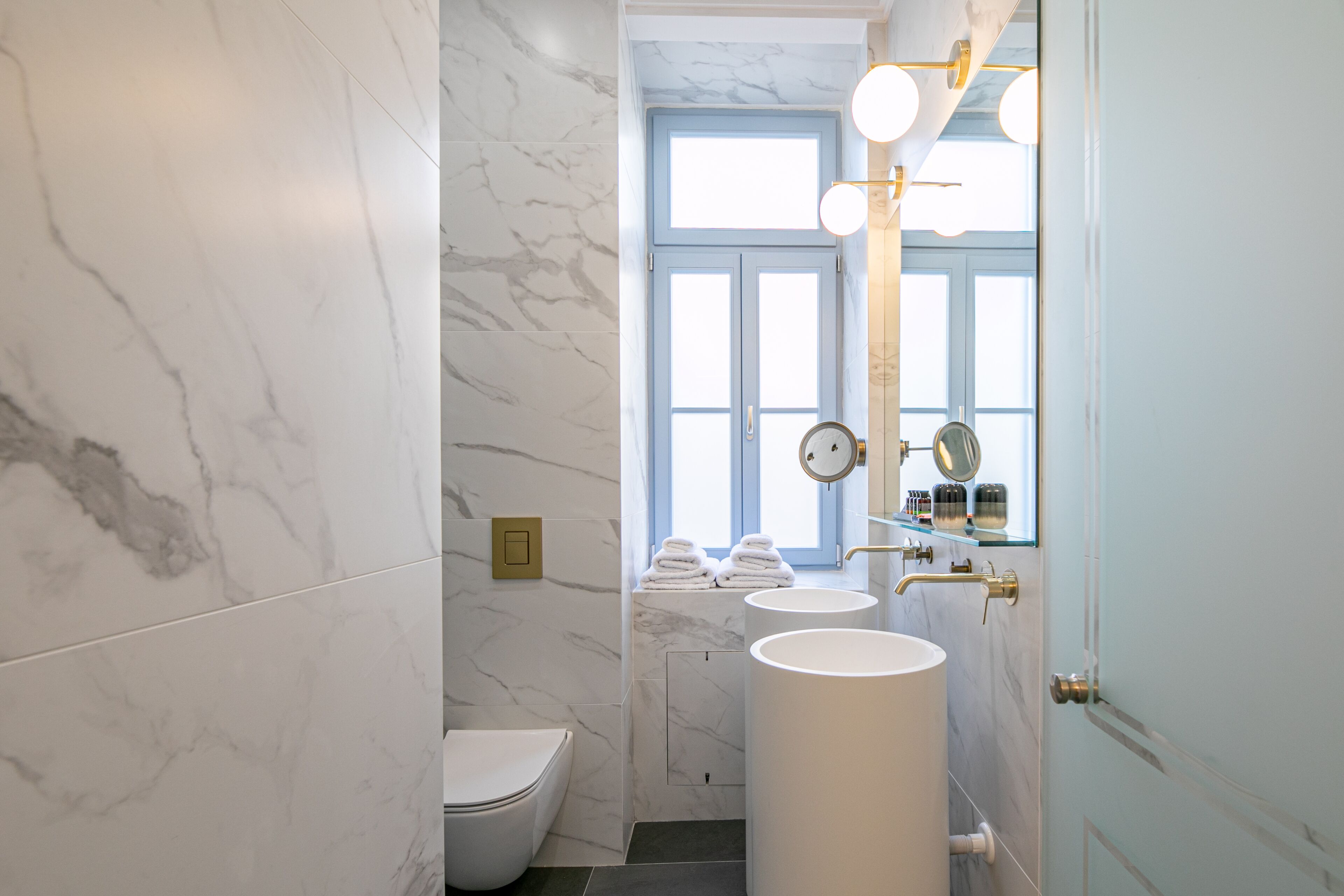 A bathroom with marble walls, a toilet, two sinks, and a window with a view.