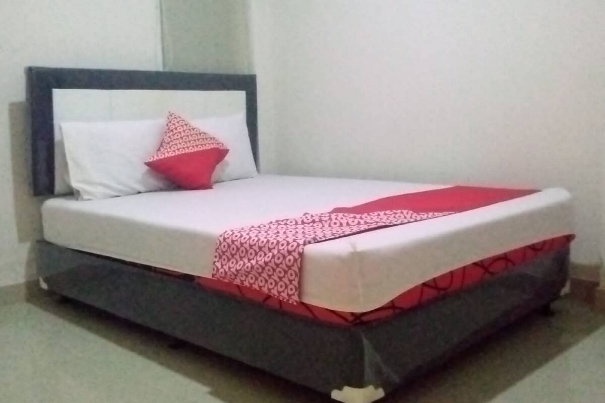 Una cama con estructura gris, ropa de cama blanca y una manta con estampado rojo y blanco.