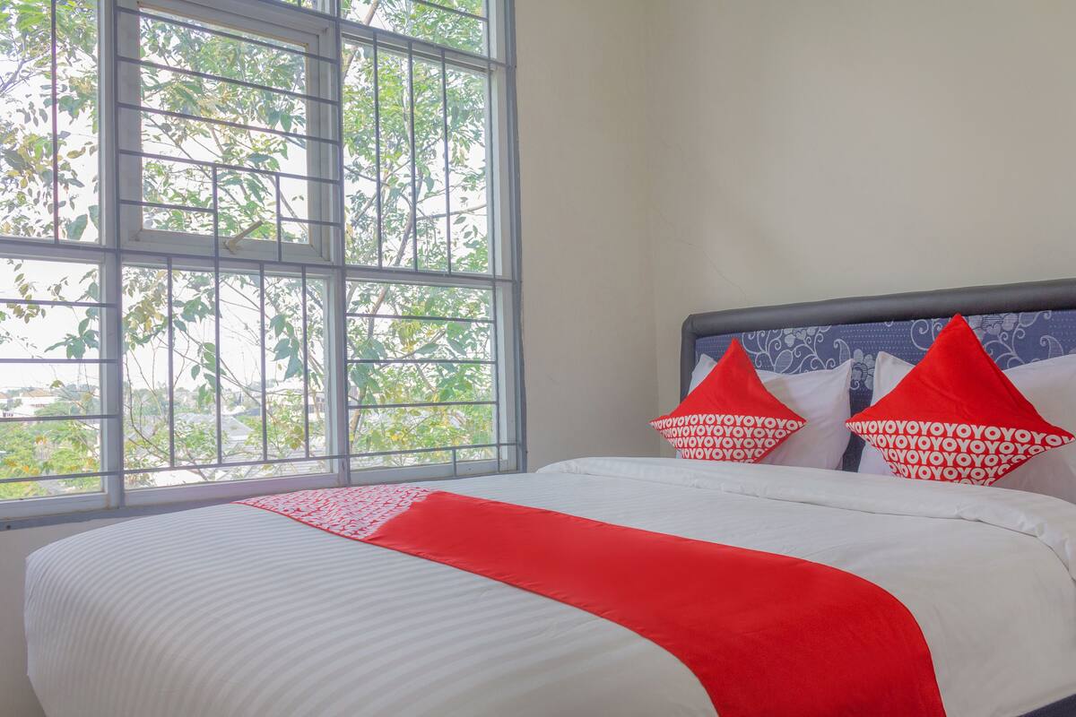 Una habitación de hotel con un ventanal grande, una cama con ropa blanca y detalles rojos, y almohadones decorativos.
