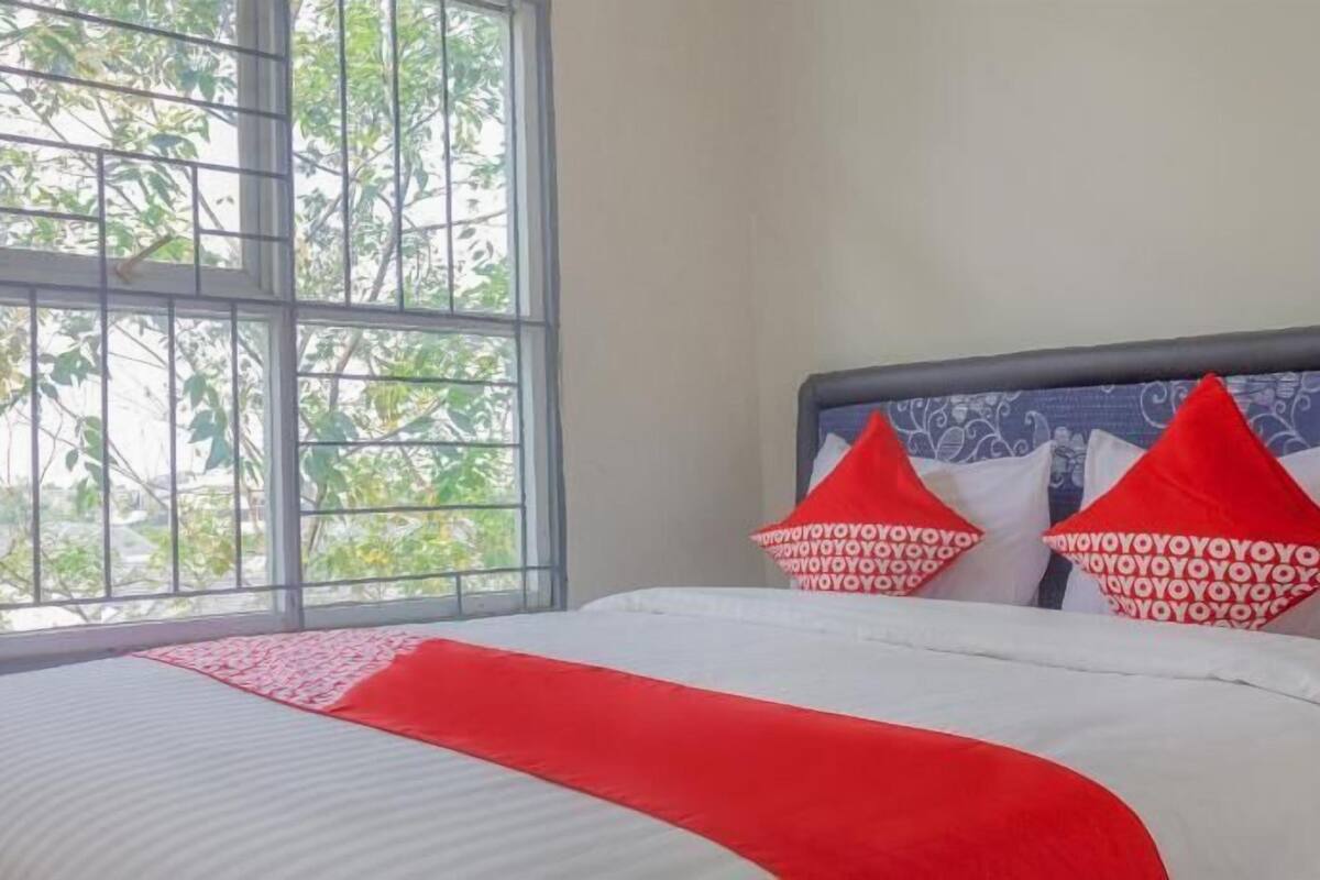 Una cama con una colcha blanca y roja, dos almohadas rojas con diseños blancos, y una ventana que da a un paisaje de árboles.