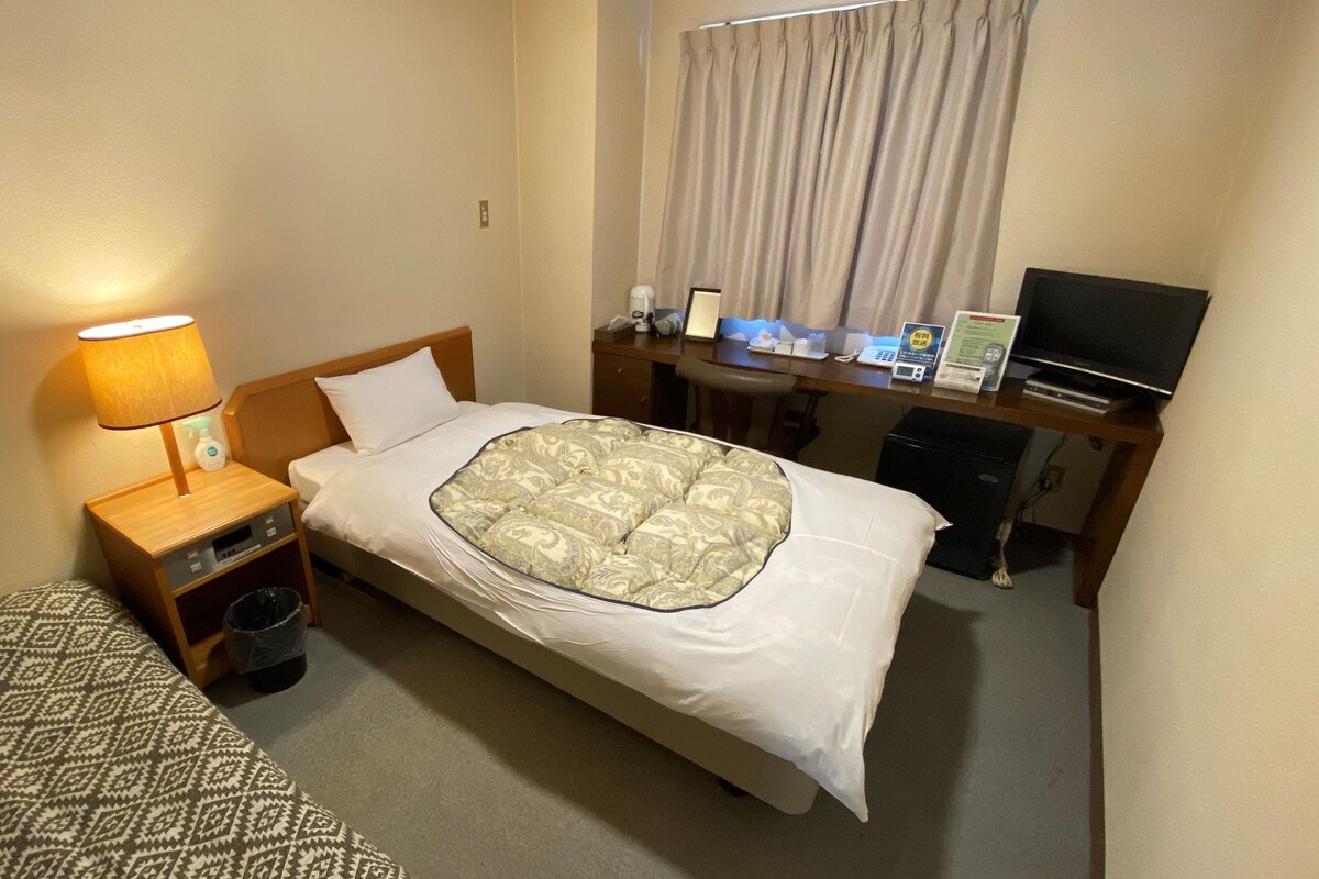 A hotel room with a bed, a bedside table with a lamp, a desk with a television, and a trash bin.