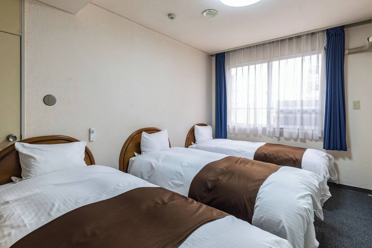 Ein Hotelzimmer mit drei Betten, einem Fenster mit blauen Vorhängen und einer Deckenleuchte.