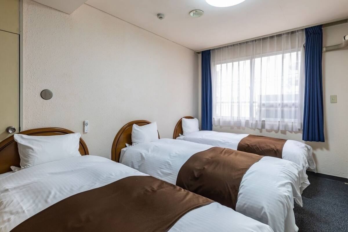 Ein Hotelzimmer mit drei Betten, einem großen Fenster mit Vorhängen und einer Deckenleuchte.