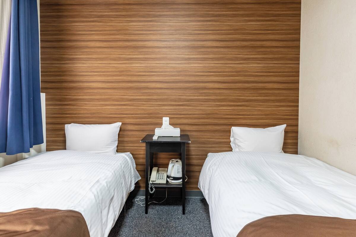 Ein Hotelzimmer mit zwei Betten, einem hölzernen Kopfteil, einem Nachttisch mit Telefon und einem blauen Vorhang.